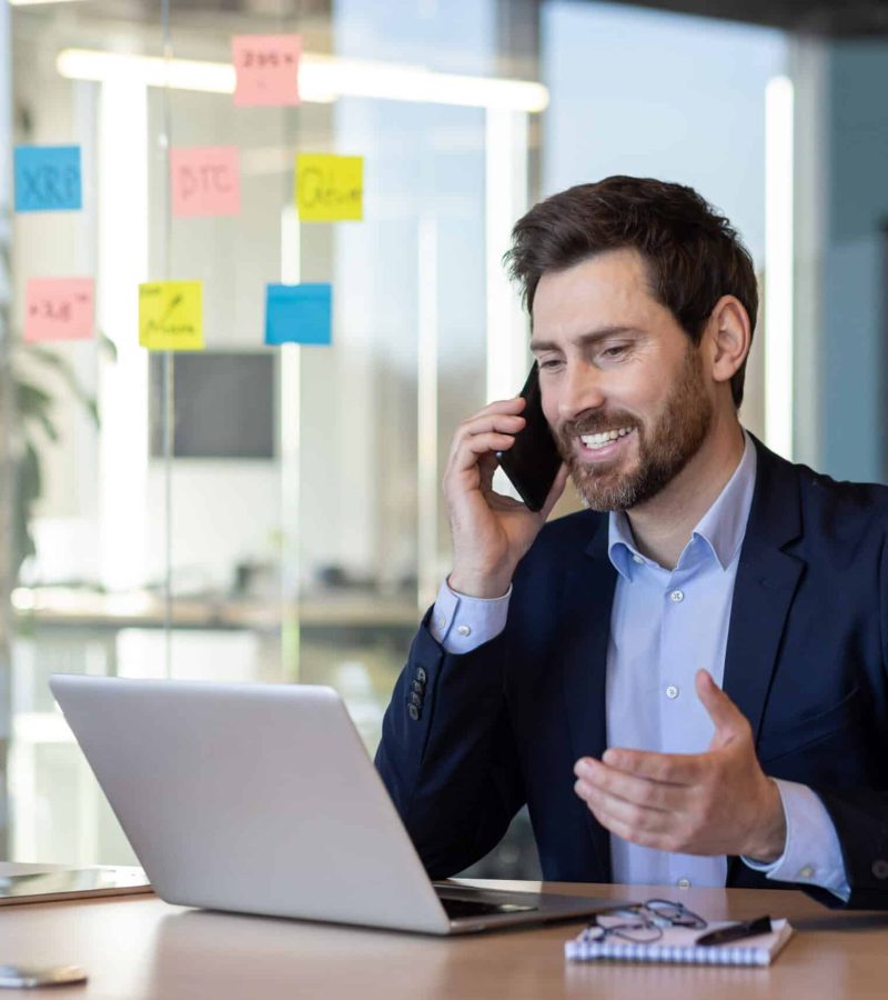 Smiling happy boss talking on the phone, businessman satisfied with the results of negotiations achieved at the workplace inside the office, mature boss in a business suit working with a laptop.
